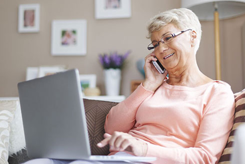 A senior woman talking on a cell phone while using a laptop.
