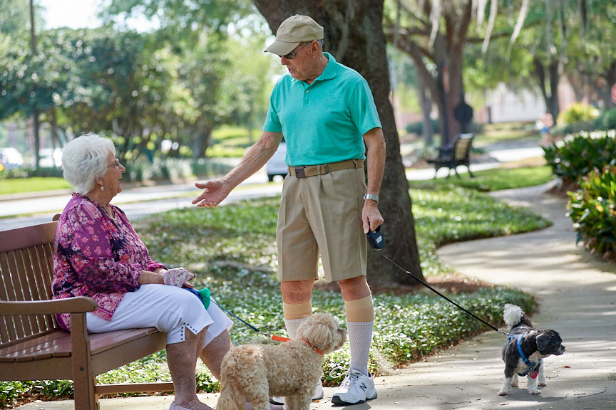 A senior man walking his small dog talks to a senior woman sitting on a park bench.