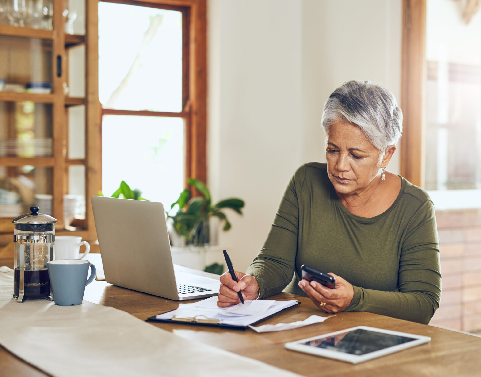 An older woman looks at paperwork and her laptop and tablet at the dining room table